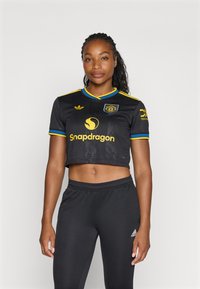 Črn skrajšan športni dres z rumenimi poudarki in okroglo logotipom. Združen z črnimi športnimi hlačami, ki imajo majhen bel logotip.