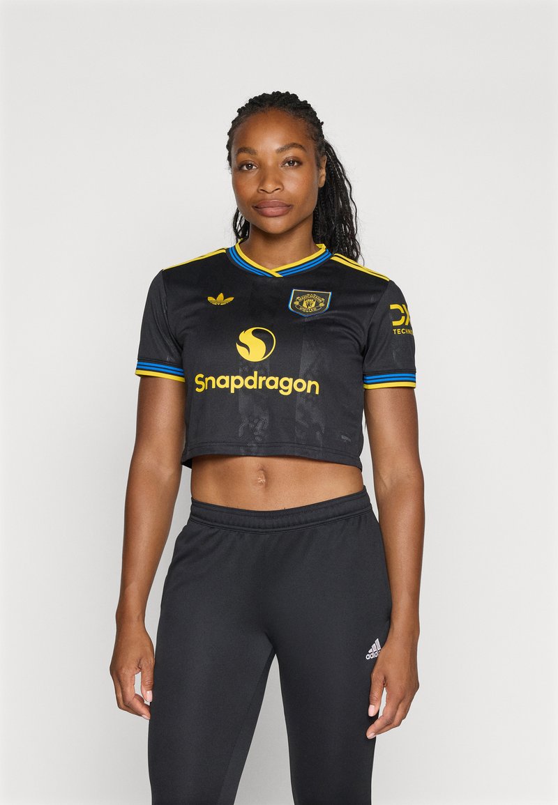 Črn skrajšan športni dres z rumenimi poudarki in okroglo logotipom. Združen z črnimi športnimi hlačami, ki imajo majhen bel logotip.