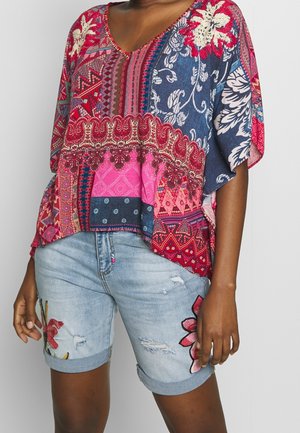 Persoon die een kleurrijke top draagt met bloemen- en geometrische patronen en lichte denimshorts met geborduurde bloemen en verweerde details.