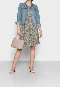 Veste en denim, robe fleurie verte et blanche, sac à main beige et sandales à talons carrés blanches. L'ensemble présente une silhouette décontractée.