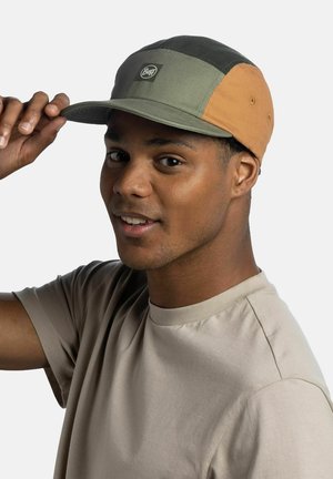 Hombre ajustándose una gorra bicolor con el logo de Buff, lleva una camiseta beige, mirando a la cámara con una leve sonrisa, sobre un fondo liso.