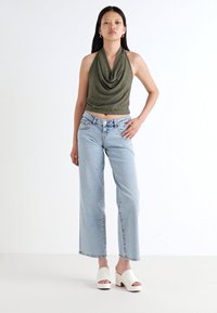 Naine seisab enesekindlalt, seljas roheline spagetisalong, heledad sinised teksad ja valged kontsakingad, ühevärvilise tausta ees.