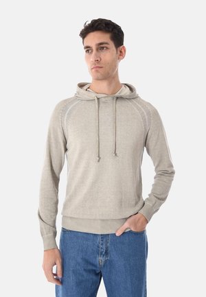 Uomo che indossa una felpa beige con cappuccio e coulisse, e jeans blu, in piedi con una mano in tasca, che guarda di lato.