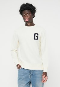 Crème kleurig sweatshirt met een ronde halslijn, geribbelde boorden en zoom. Bevat een marineblauwe patch met de letter "G" op de linker borst. Draag het met een blauwe spijkerbroek.