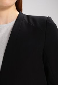 Černé sako s výraznými rameny, hladkou tkaninou a klopovým límcem s výstřihem, vrstvené přes světle šedý texturovaný svetr.