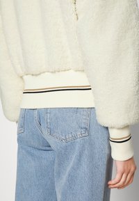 Person i ljusblå denimjeans och en vit fleecejacka med randiga ribbade manchettar och midja, bakvy.