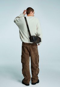 Nadrozmerný krémový rebrovaný sveter, hnedé cargo nohavice s viacerými vreckami, čierna crossbody taška, stojaci proti svetlému pozadiu.