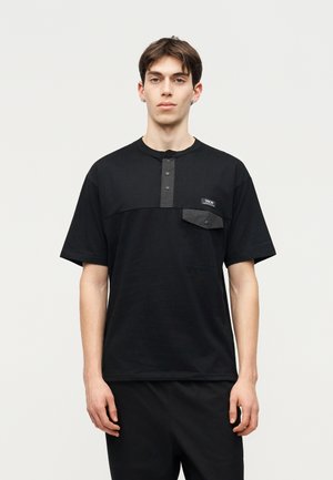 Nuori mies mustassa lyhythihkaisessa paidassa, jossa on napillinen rinta-aukkko ja rintatasku, seisoo yksinkertaisen vaalean taustan edessä.