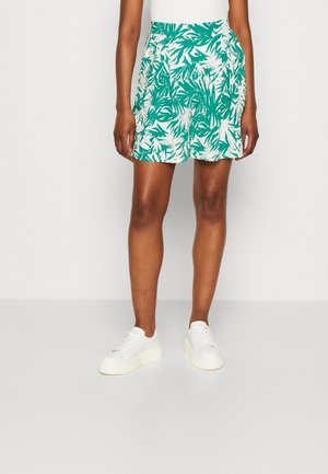 Kvinde iført grønne og hvide shorts med bladmønster og hvide sneakers, stående mod en ensartet lys grå baggrund.