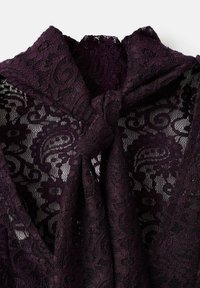 Tela de encaje de un profundo color púrpura con intrincados patrones de flores y espirales, con un detalle en el escote atado. Acabado suave y texturizado.