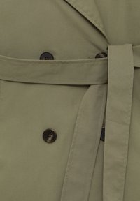 Tkanina oliwkowego płaszcza trencherowego o matowej fakturze, z dwoma dużymi brązowymi guzikami, przewiązanym paskiem oraz klasycznym kołnierzem.