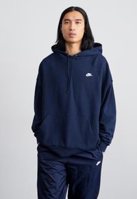 Ναυτικό μπλε φούτερ με τσέπη μπροστά, κουκούλα με κορδόνια και λευκό λογότυπο Nike στο στήθος, σε συνδυασμό με αντίστοιχες ναυτικές φόρμες.