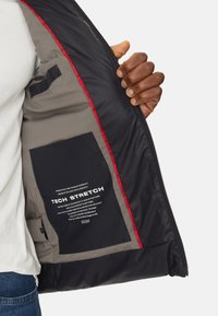 La chaqueta acolchada negra presenta un interior con forro gris, un bolsillo etiquetado como Tech Stretch y detalles de costura roja a lo largo del zipper.