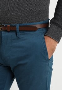 Homme portant un pantalon bleu avec une ceinture en cuir marron et une chemise à manches longues gris foncé texturée, la main reposant dans la poche.