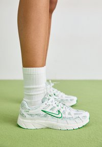 Nike P-6000 tenisice u bijeloj boji s zelenim naglascima, s teksturiranim dizajnom, mrežastom gornjom plohom i uzorkovanim preklopima. Nosene s bijelim rebrastim čarapama.