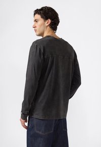 Junger Mann mit dunklem, lockigem Haar, der ein ausgeblichenes schwarzes Langarmshirt und dunkelblaue Jeans trägt, und nach links auf einem weißen Hintergrund schaut.