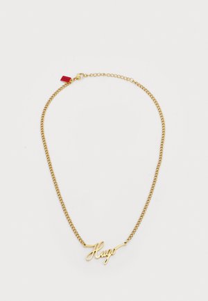 Gouden ketting met de naam "Hugo" in cursief schrift en een klein rood labeltje bij de sluiting op een witte achtergrond.