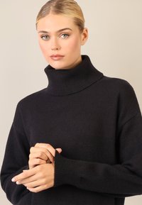 Schwarzer, übergroßer Pullover mit hohem, geripptem Kragen und Ärmeln bis zur Handgelenke. Weiche Textur mit weiten Armausschnitten und lockerer Passform.