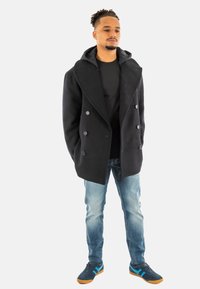 Abrigo negro tipo peacoat con grandes solapas y botones de doble breasted, usado sobre una camiseta negra, combinado con jeans azul claro y zapatillas azules con rayas.
