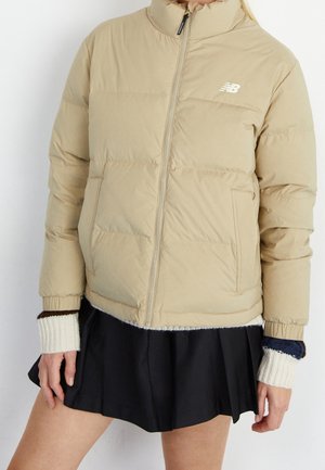 Veste matelassée beige New Balance avec fermeture éclair frontale portée sur une jupe noire plissée et des manches tricotées superposées crème et bleu marine.