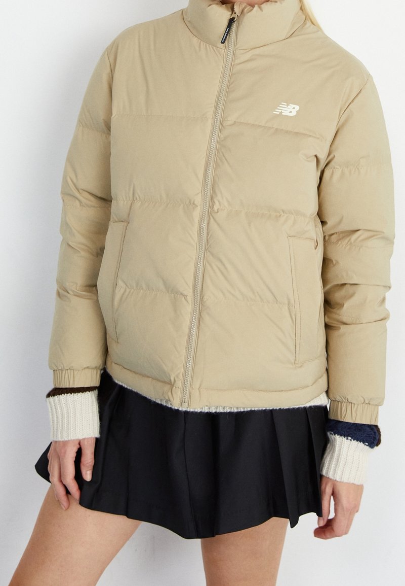 Veste matelassée beige New Balance avec fermeture éclair frontale portée sur une jupe noire plissée et des manches tricotées superposées crème et bleu marine.