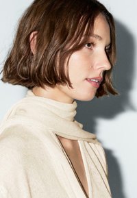 Beige knit scarf wrapped around the neck, draped over a light, textured garment. Short brown hair, soft features, and a neutral background.
