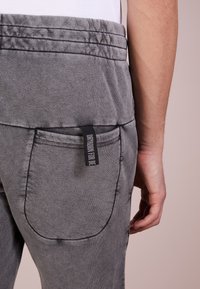 Osoba v šedých teplácích s kapsou na zadní straně a černou nálepkou s nápisem "DRYWORN FOR BE" viditelnou v blízkosti pasu.
