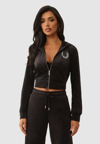 Haut de sweatshirt noir à zip court en tissu doux, doté de poches avant et d'un détail décoratif argenté sur le côté gauche. Associé à un pantalon assorti.