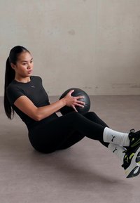 Black fitted athletic top and leggings on a person performing an exercise with a black medicine ball, wearing white socks and black sneakers.