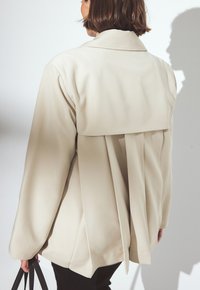 Beige tailored jacket with long sleeves, a back pleat design, and a wide collar. Material appears smooth with a slight sheen.
