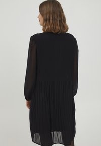 Schwarzes plissee Kleid mit transparenten langen Ärmeln, knöchellang, mit hohem Ausschnitt und gerafftem Taillenbereich. Glatte Textur und fließendes Design.
