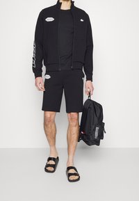 Homme portant une veste zippée noire Lacoste, un short noir, des sandales noires, tenant un sac à dos noir avec des détails graphiques blancs.
