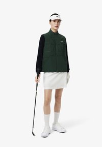 Groene gewatteerde vest met ritssluiting, voorzien van een logo, gecombineerd met een witte rok, witte sokken en witte sneakers, terwijl hij een golfclub vasthoudt.