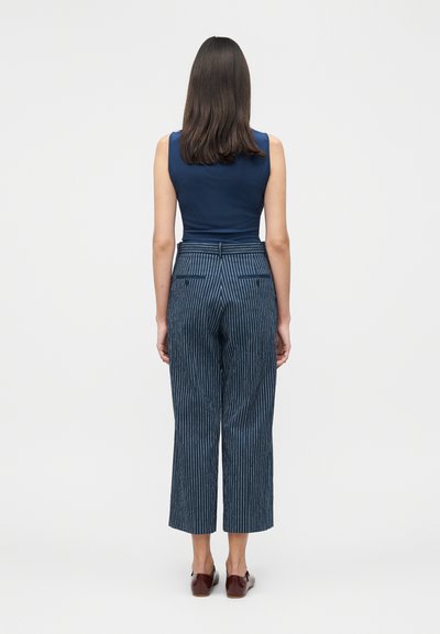 Γυναίκα με καστανά μαλλιά μέσου μήκους, φορεί ένα σκούρο μπλε αμάνικο τοπ, μπλε φαρδιά παντελόνια με λεπτές ρίγες και καφέ παπούτσια, βρίσκεται με την πλάτη σε λευκό φόντο.