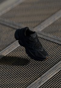 Scarpa sportiva nera con tomaia in rete, motivi testurizzati e suola flessibile. Include lacci neri e un logo discreto sul lato.