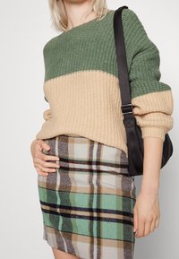 Gebreide trui met een groen en beige kleurblokontwerp, gecombineerd met een geruite rok in tinten bruin, teal en zwart. Zwarte tas.