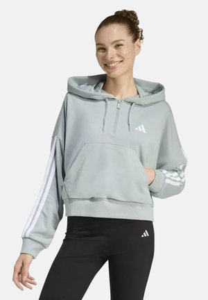 Smiling woman with curly hair in a light blue hoodie with white stripes and black leggings, standing against a plain background.