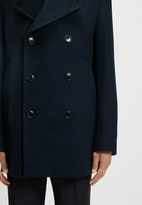 Cappotto doppiopetto in lana navy con grandi bottoni neri, colletto a punta e texture liscia, abbinato a pantaloni scuri e texture.