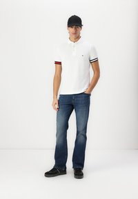 Hvit polo skjorte med svarte og røde stripete mansjetter, kombinert med blå jeans og svarte sko. Modellen har på seg en svart caps, stående mot en nøytral bakgrunn.