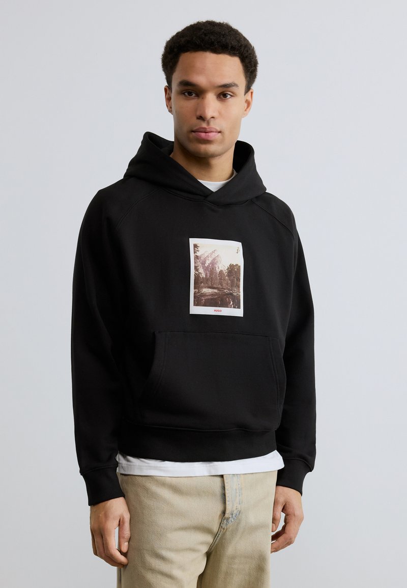 Νεαρός άνδρας που φορεί μαύρο φούτερ με κουκούλα με φωτογραφικό τύπωμα βουνού και δάσους, μπεζ παντελόνι, στέκεται μπροστά σε απλό ανοιχτό γκρι φόντο.