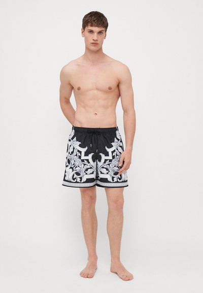 Shorts de bain noirs avec un motif orné blanc, dotés d'une taille élastique et d'un cordon de serrage. Le tissu semble léger et lisse.