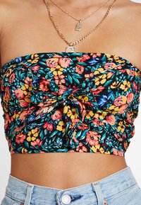 Femme portant un haut sans manches à fleurs avec un nœud à l'avant, un jean taille haute bleu clair, des colliers en chaîne dorée avec des pendentifs rectangulaires.