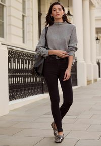 Pilkas megztinis su aukštu apykakliu, derinamas su juodais aptemptais džinsais. Dėvimi blizgūs plokšti batai ir didelė juoda rankinė.