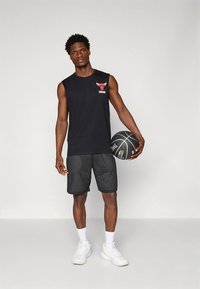 Melna krekla bez piedurknēm ar sarkanu Čikāgas Bulļļu logotipu, melnas sieta šorti un baltas sporta kurpes. Figūra tur basketbola bumbu.