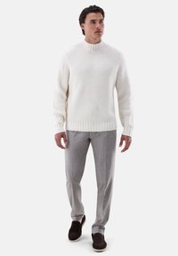 Weißer strukturierter Strickpullover mit hohem Kragen, kombiniert mit hellgrauen maßgeschneiderten Hosen und dunkelbraunen Schuhen.