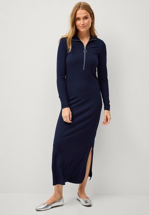 Mørkeblå, ribbet kjole med lange ærmer, halv lynlås krave, sideslidser og tætsiddende silhuet, parret med sølvfarvede ballet flats.