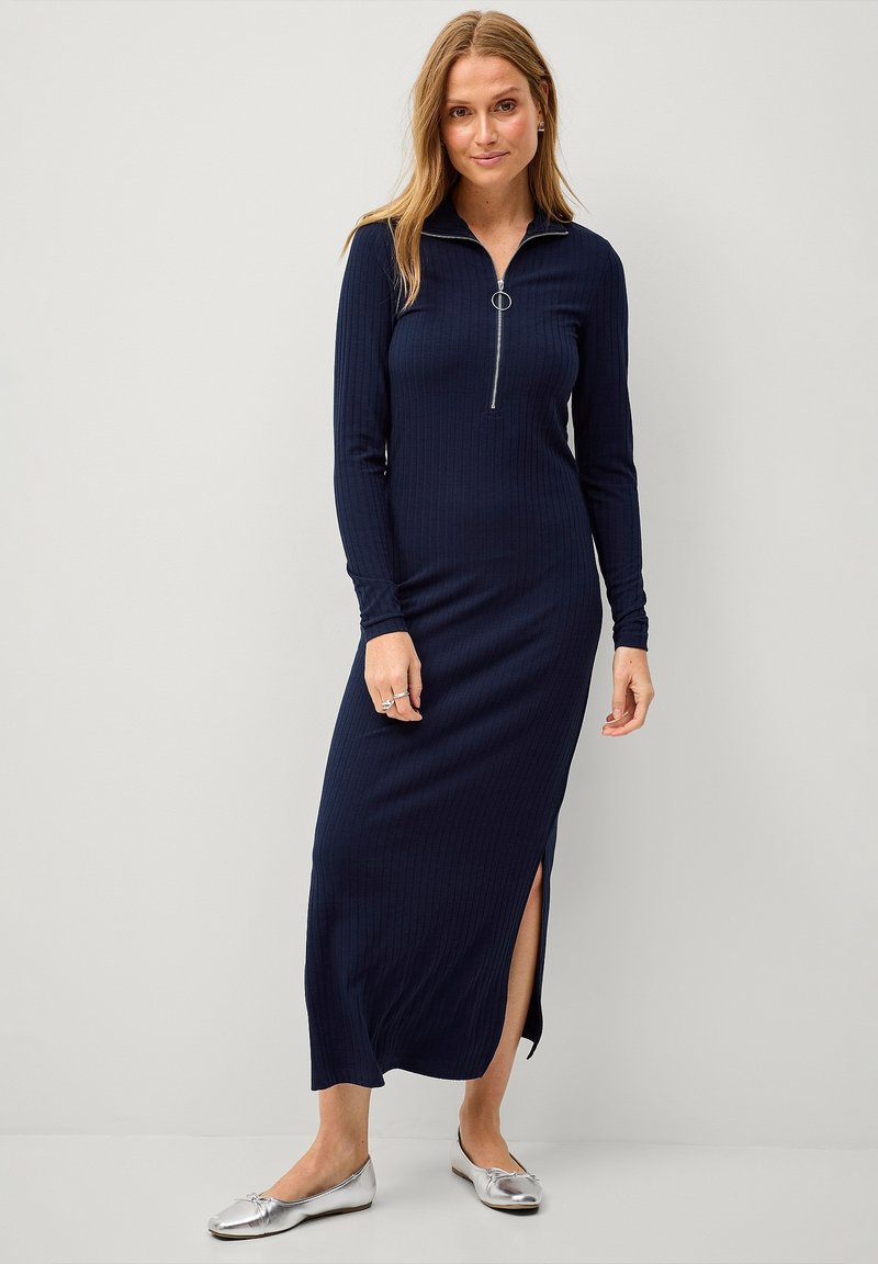 Marineblaues, geripptes Langarmkleid mit einem halben Reißverschluss-Kragen, seitlichen Schlitzen und einer taillierten Silhouette, kombiniert mit silbernen Ballerinas.