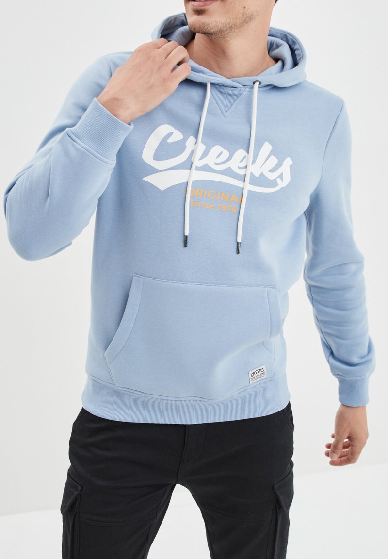 Felpa leggera blu chiaro con tasca frontale, cappuccio con laccetti e logo "Creeks" bianco con testo arancione. Realizzata in tessuto morbido con polsini a coste.