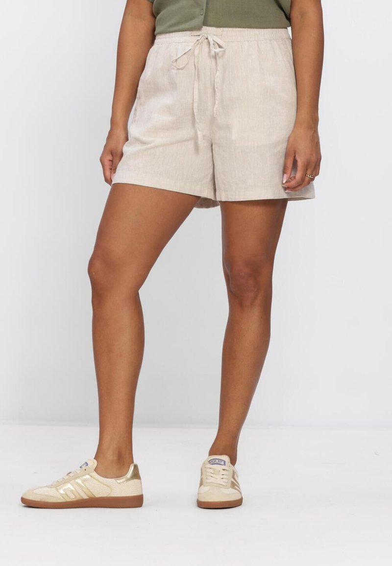Personne portant un short beige avec cordon de serrage et des baskets crème, debout devant un fond clair uni.