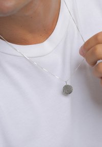 Ezüst medál nyaklánc, texturált, kör alakú medállal, finom láncon, sima fehér póló fölött viselve.